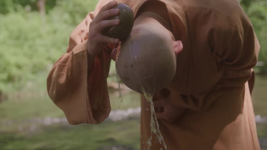 A person in an orange robe is using a half coconut shell to pour water over their bald head the water streams down the persons head and neck