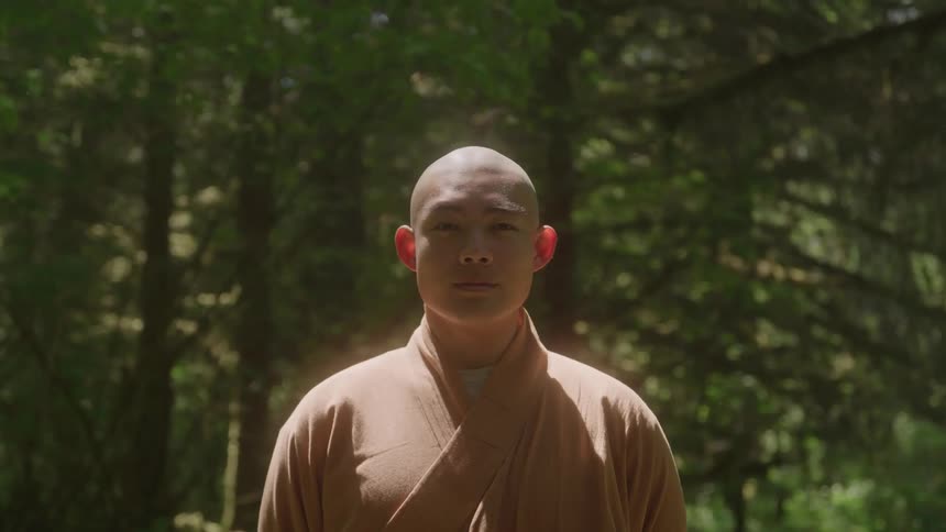 A tibetan monk with a shaved head and wearing a light brown robe is standing in a lush forest with tall trees and a green underbrush looking directly at the camera