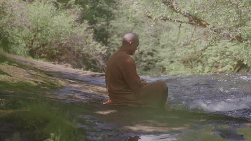 A monk with a shaved head is seated peacefully on a rock next to a flowing river or stream the monk dressed in orange robes is surrounded by lush greenery and appears to be meditating or contemplating