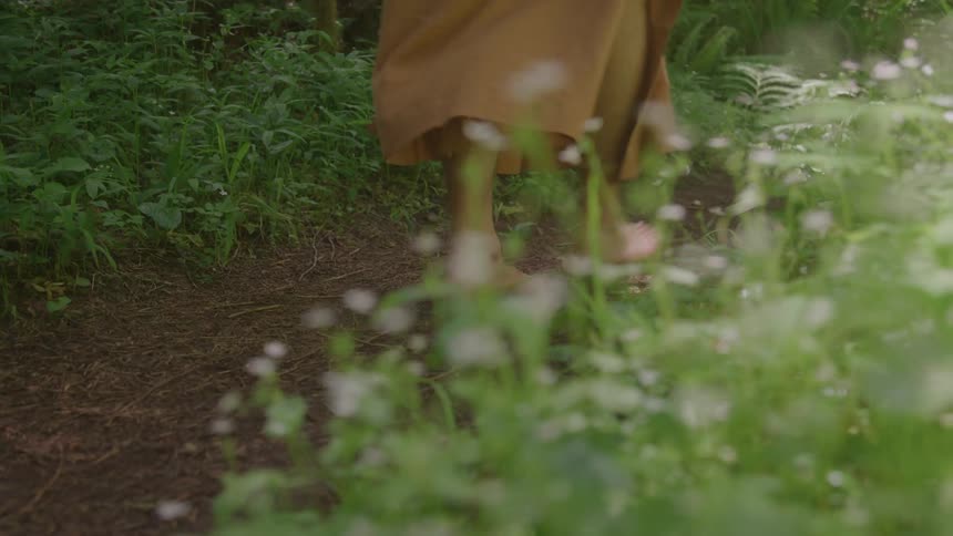 The video shows a path through a lush green forest or garden a persons bare feet are seen walking along the dirt path their long tan garment trailing behind them