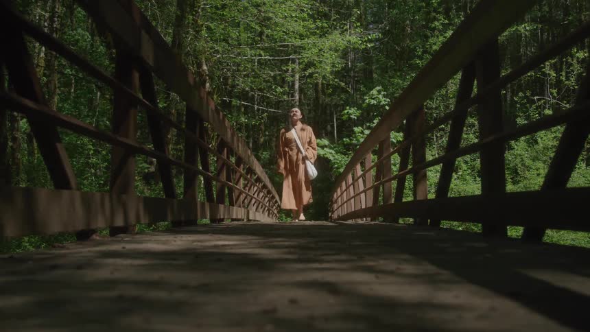From a very low angle on the floor of a wooden bridge a barefoot monk walks from the far end approaches the camera and steps over it