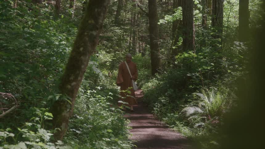 The camera reveals from behind a tree to show a monk in long robes walking towards the camera down a dirt path in a dense forest