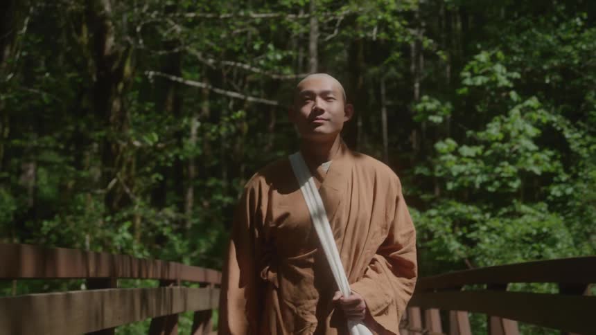 A monk in traditional robes walks toward the camera across a wooden bridge looking around at the sunlit forest surroundings with a peaceful expression