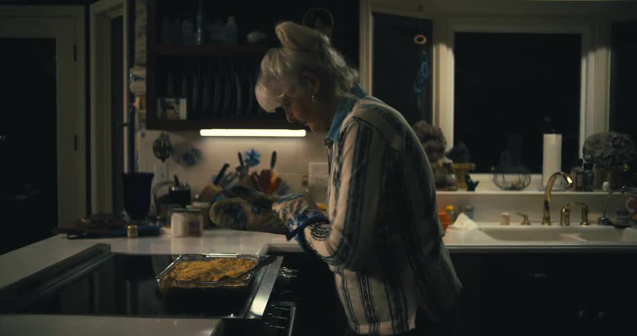 A woman with her hair tied up uses oven mitts to carefully remove a baking dish from an oven in a kitchen