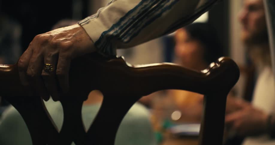 A close angled view shows a persons hand with wrinkled skin and a gold ring resting on the curved wooden back of a chair