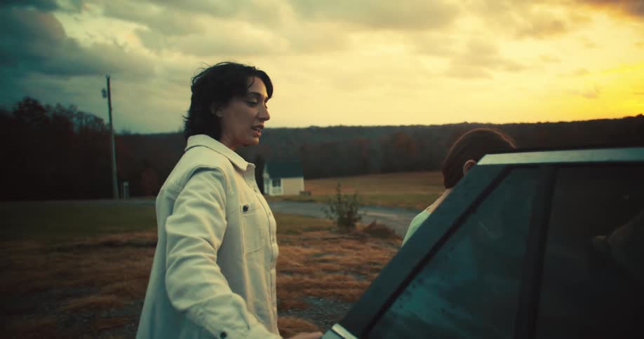 An adult woman and a girl exit a silver vehicle and walk away from it at sunset towards a white house