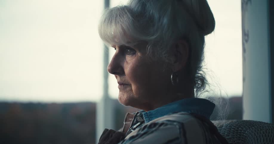 The video shows a closeup of an older woman with gray hair styled in a bun she is looking out a window with a pensive expression and the background is slightly out of focus