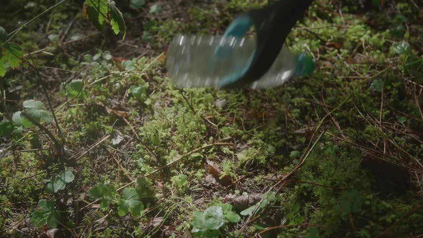A green grabbing tool reaches down and picks up a clear plastic bottle that is lying on the ground covered in green moss and small plants