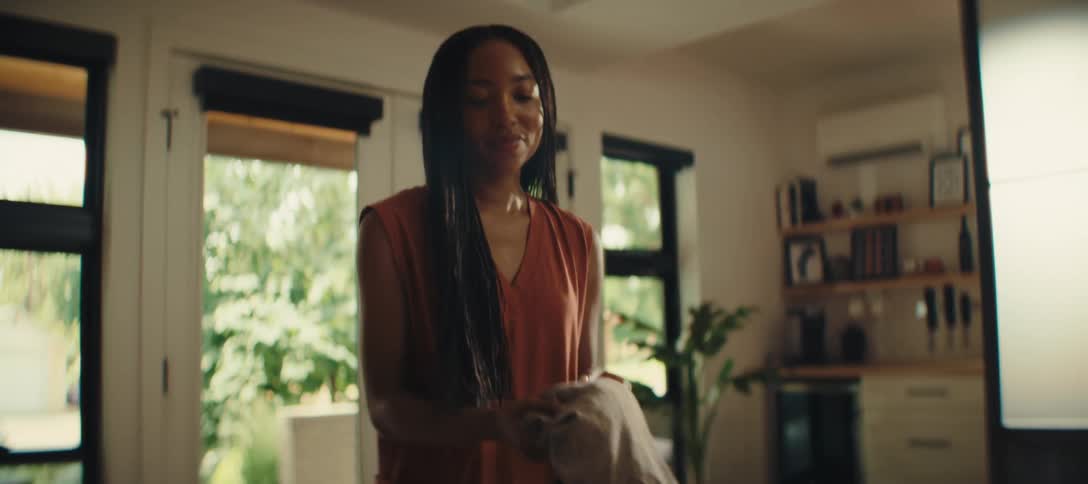 A woman with long dark hair wearing a loose fitting rust colored sleeveless top is standing in a bright room with a large suitcase open on a bed packing clothing into the case