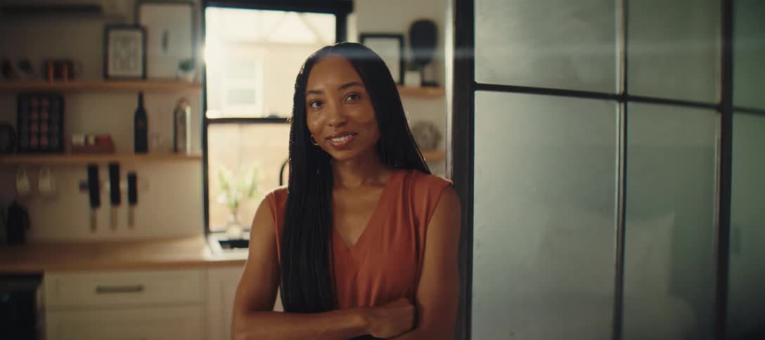 A smiling woman with long braided hair stands in a kitchen with her arms crossed her head is tilted slightly to the side as she looks at the camera