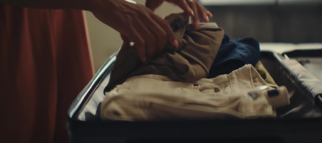 A young black woman with long braided hair arranges clothes into a suitcase on her bed at midday