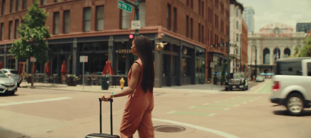 A woman with long braids wearing an orange sleeveless jumpsuit pulls a black suitcase behind her as she walks across a street in a city
