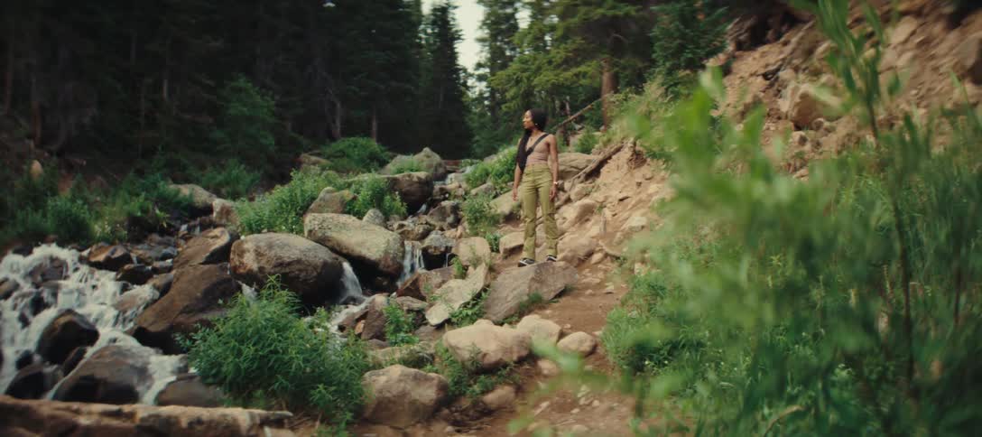 A young woman hiking in the forest climbs down a rocky path with a stream flowing below at midday