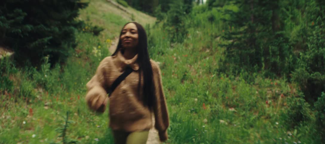 A young woman is walking down a dirt path in a grassy area the path is surrounded by green plants and some red flowers