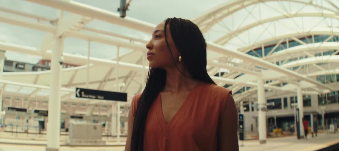 The video shows a woman with long black braids standing on a train platform she pulls a black suitcase behind her and looks up with a slight smile as she checks a gold watch on her wrist