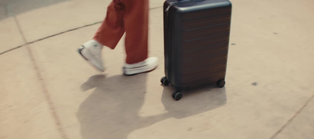 A woman with long braids and a sleeveless orange outfit rolls a black suitcase with a retractable handle along a sidewalk in the city