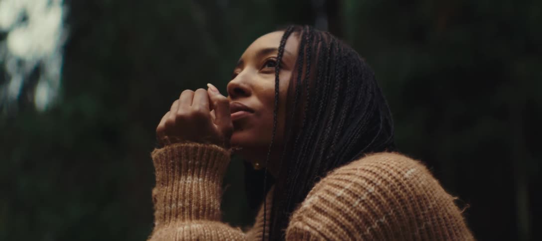 A woman with braids and a brown knit sweater raises her hands to her chin closes her eyes and looks up