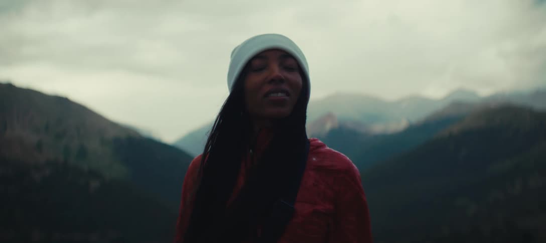 The video shows a woman standing in a mountainous forested area she is wearing a red jacket and a lightcolored beanie she turns to face the camera and then smiles