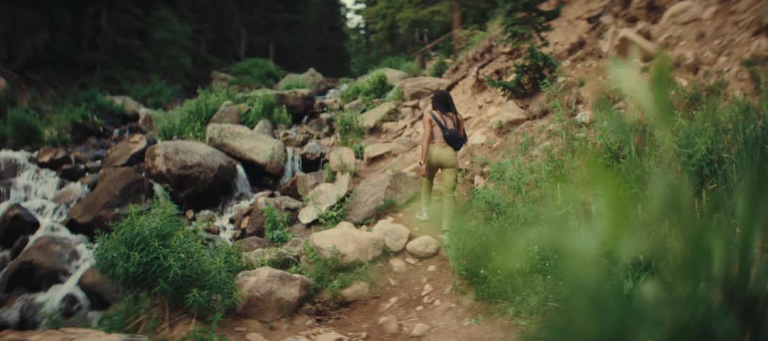 A young woman wearing light green pants and a black tank top with a black crossbody bag is hiking up a rocky trail next to a small waterfall in a forest