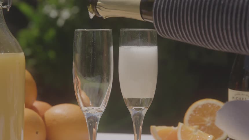 Champagne is poured into two flute glasses already containing some orange juice to create mimosas on an outdoor table surrounded by oranges and orange slices