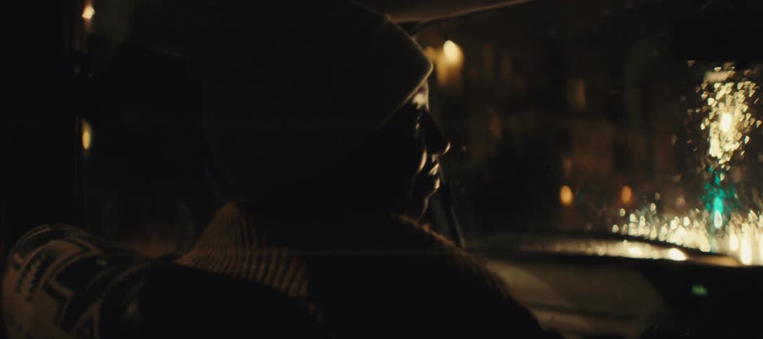 A person with a light colored beanie and a brown sweater sits in the passenger seat of a car at night facing left and looking out a rain streaked window at the blurred lights of a city street
