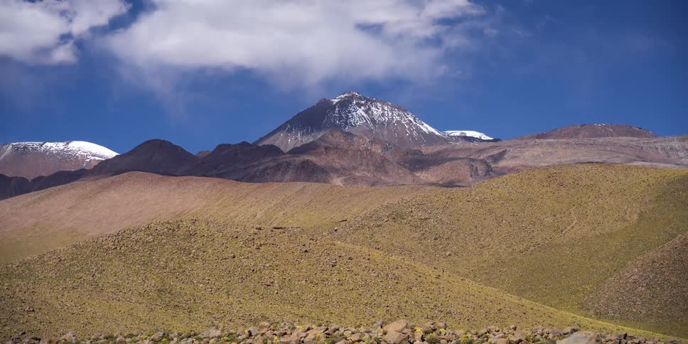 In a timelapse the shadows of clouds sweep quickly across the dry rolling hills of a desert landscape with a snowcapped mountain in the background