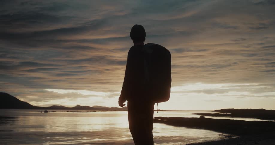 A person with a backpack and a beanie is standing on a beach at sunset silhouetted against a golden and cloudy sky and a calm body of water with distant land masses
