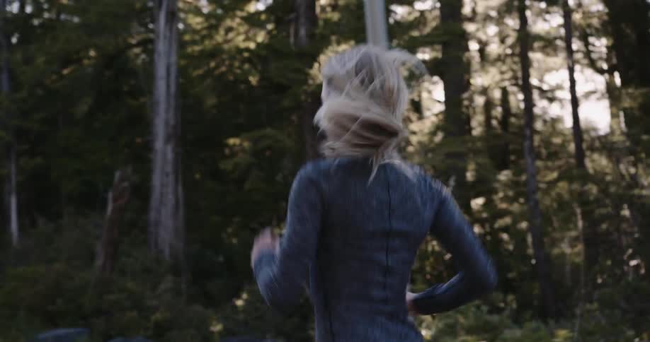A young woman with blonde hair wearing activewear runs through the woods at midday