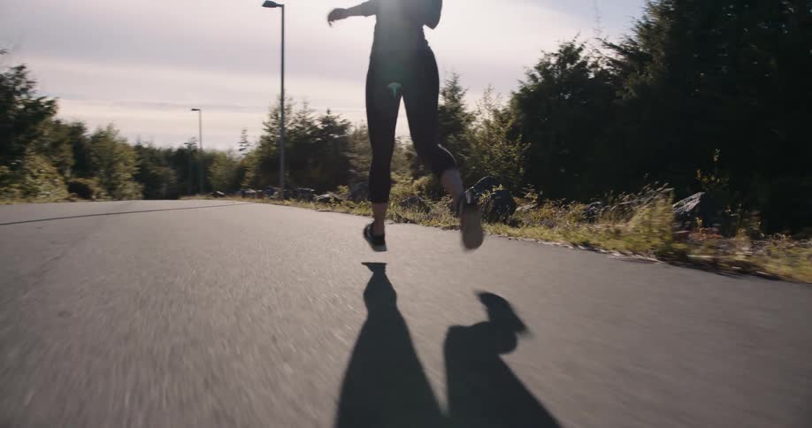 A young woman wearing active pants runs along a lonely road lined with grasses and trees on either side at midday