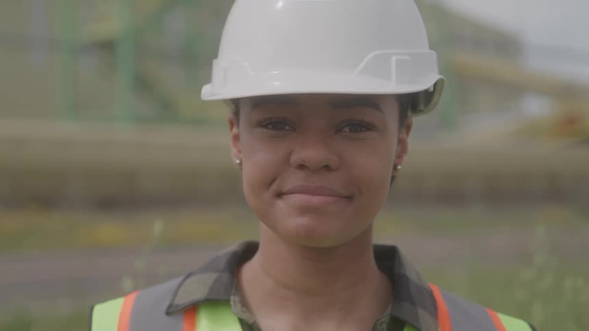 A young female construction worker stands outdoors on an open field at midday wearing a white hard hat and green vest smiling