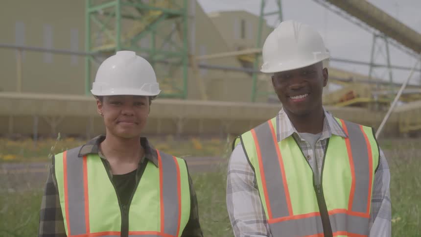 Based on the images from the video a man and a woman are standing sidebyside outdoors wearing hard hats and safety vests the man who is on the right is smiling at the camera while the woman looks straight ahead