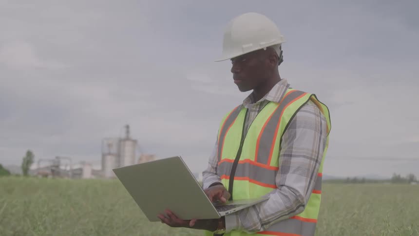 Engineer wearing a reflective vest and helmet using a laptop in an open field