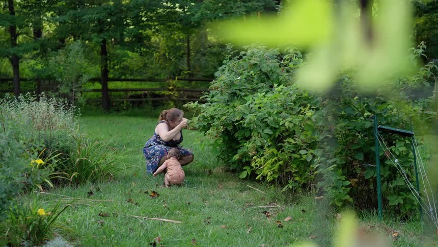 A woman crouching in a garden picking fruits from a plant with a baby beside her also picking fruits