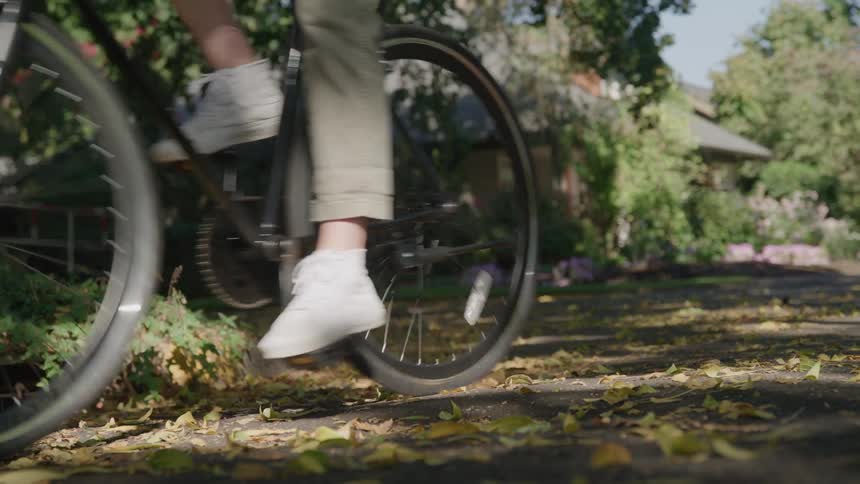 A person on a bicycle with the focus on the wheels and legs as they begin to pedal and ride away down a leafy suburban street