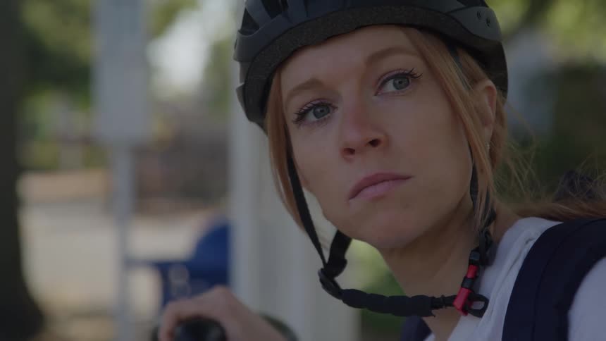 A woman with light colored hair and blue eyes wearing a black bicycle helmet is looking to the left with a serious expression with a bike handle visible in the foreground