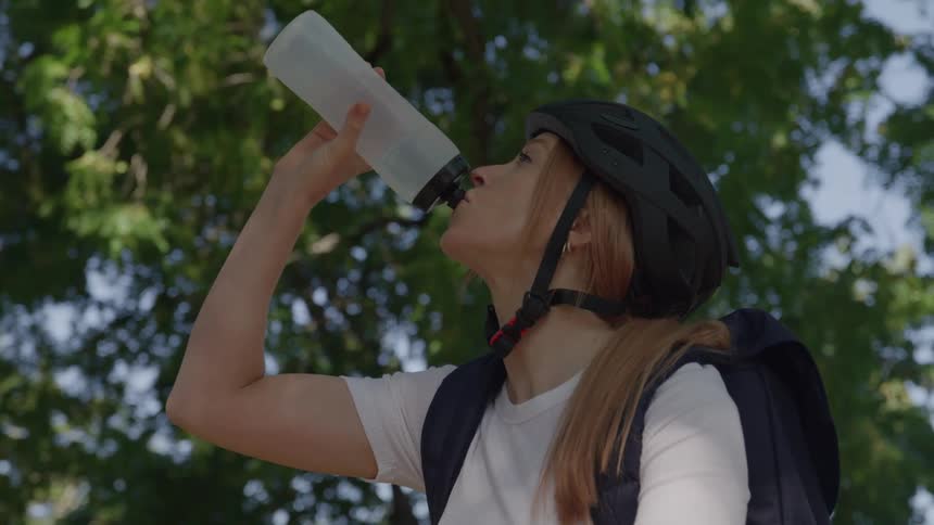 Based on the images from the video a young woman with long blonde hair wearing a white tshirt a backpack and a black cycling helmet is shown she is taking a break to drink from a clear water bottle the scene takes place outdoors with a backdrop of