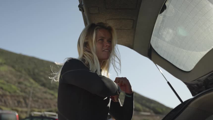 A woman standing at the back of her car looks like she is putting on deodorant