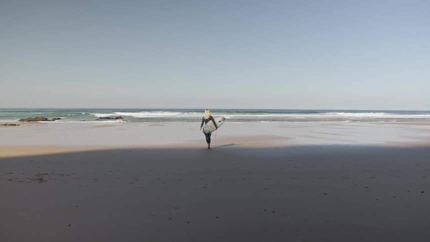 Female surfer walking on sand carrying surfboard towards the ocean