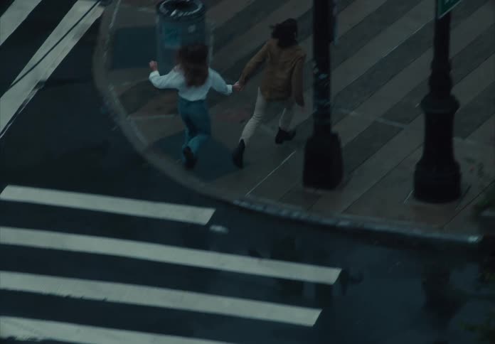 The video filmed from an elevated angle shows a young couple holding hands and running across a wet crosswalk the shot pans to follow them as they reach the sidewalk passing a streetlamp and a bright red umbrella on the ground