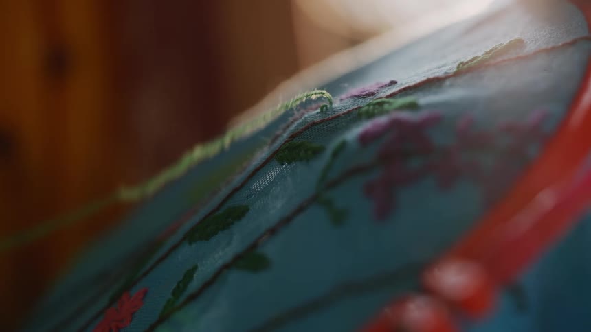 A close shallow focus view shows a persons thumb and index finger holding a needle to pull a green thread through a blue fabric with a pink and green floral design
