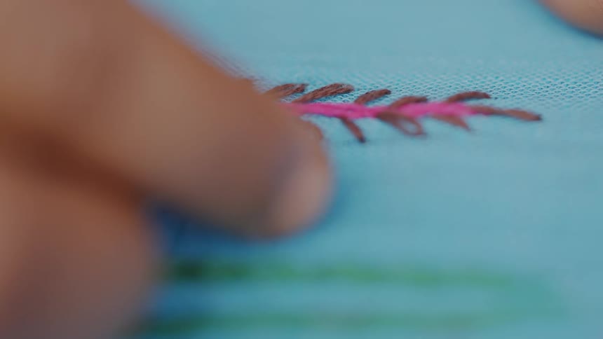 Based on the images from the video a closeup shot of a persons hands performing an intricate embroidery or sewing technique on a piece of cloth is shown a needle with a white thread is seen pulling through a blue material and joining a pink and brown