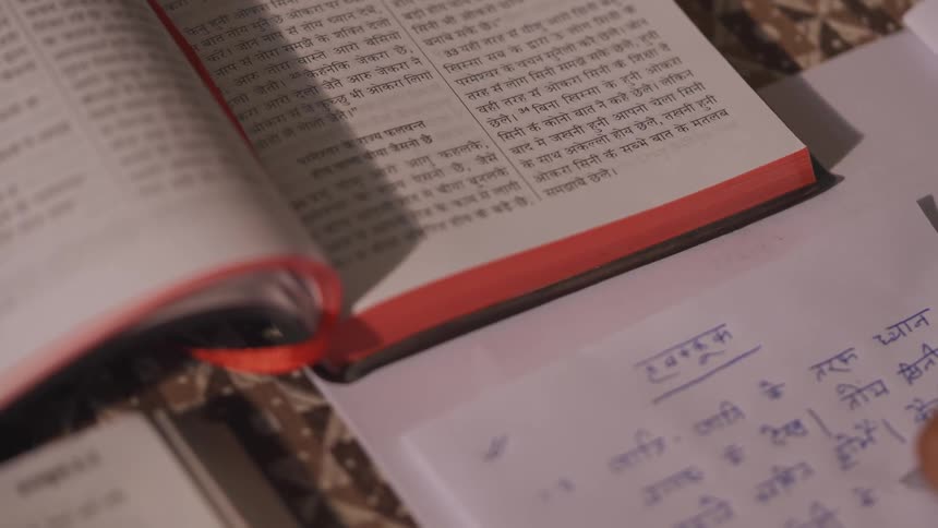 A large open book with a red cover and a red ribbon bookmark is lying on a table next to a persons hand who is using a blue pen to copy hebrew text from the book onto a white sheet of paper