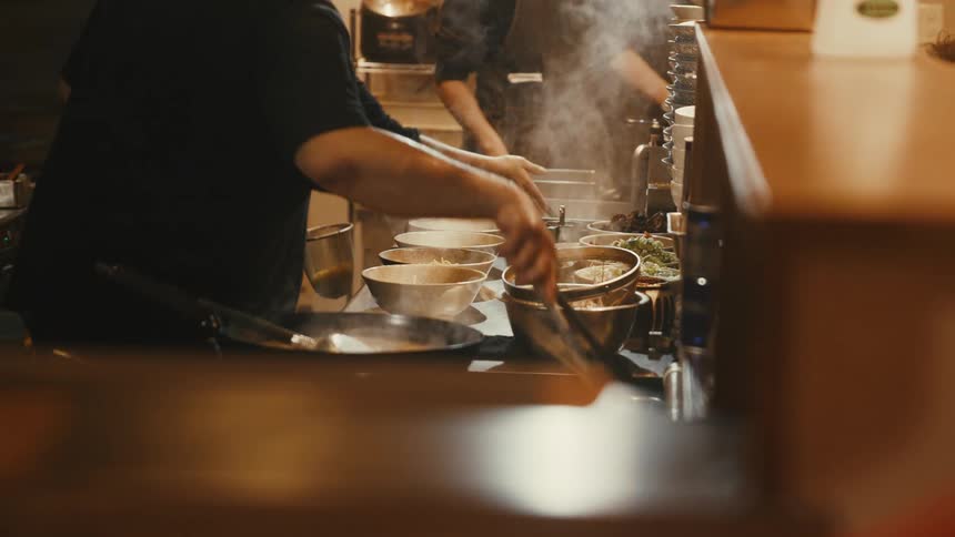 Cooks prepare hot dishes in a professional kitchen with bowls lined up on the counter