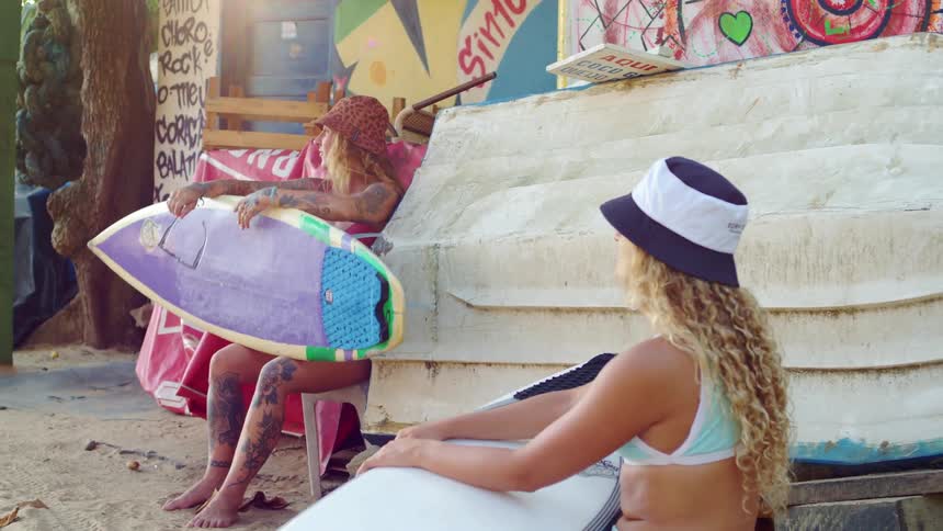 Two women sitting on the beach in front of a cabin holding surfboards on their laps while talking to each other