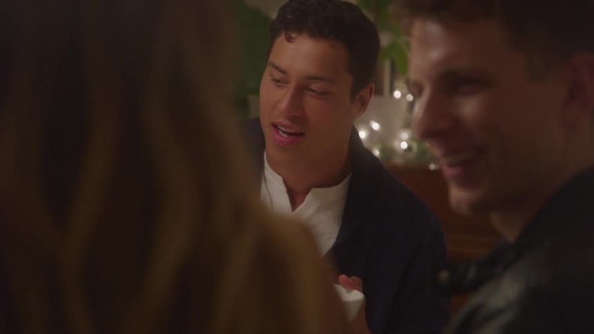 A man with short dark hair is sitting at a table with two other people smiling as he looks down at something in his hands while wearing a dark jacket over a white shirt