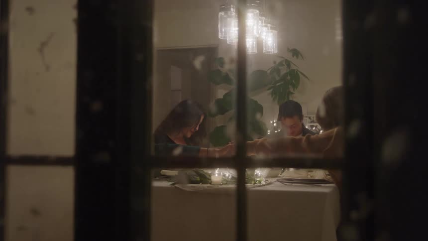 A group of friends is sitting at a candle lit dinner table as he raises their hands in prayer all visible through the blurry glass panes of a window at night with snow falling