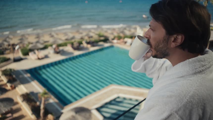 A man in a bathrobe drinks coffee at a poolside resort