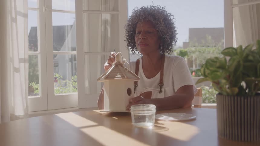 Woman paints a wooden birdhouse while sitting at a table near the window