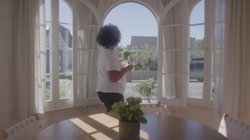 Woman stands holding mug and looking outside of large window