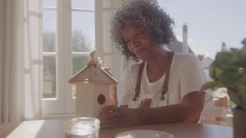 Elderly woman smiles while holding a birdhouse on a sunlit table